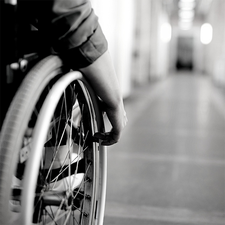 A monochrome image of a person in a wheelchair. Their back is to the camera and they are looking down an empty corridor.