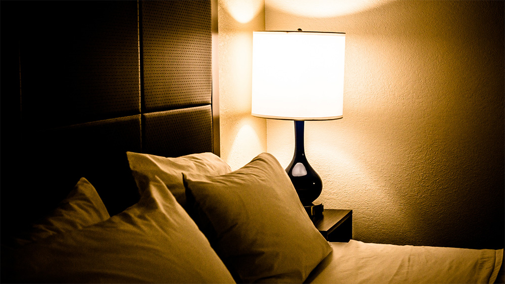 A photograph of a double bed at night time. On the night stand next to the bed is a lamp. The photo is sepia toned.