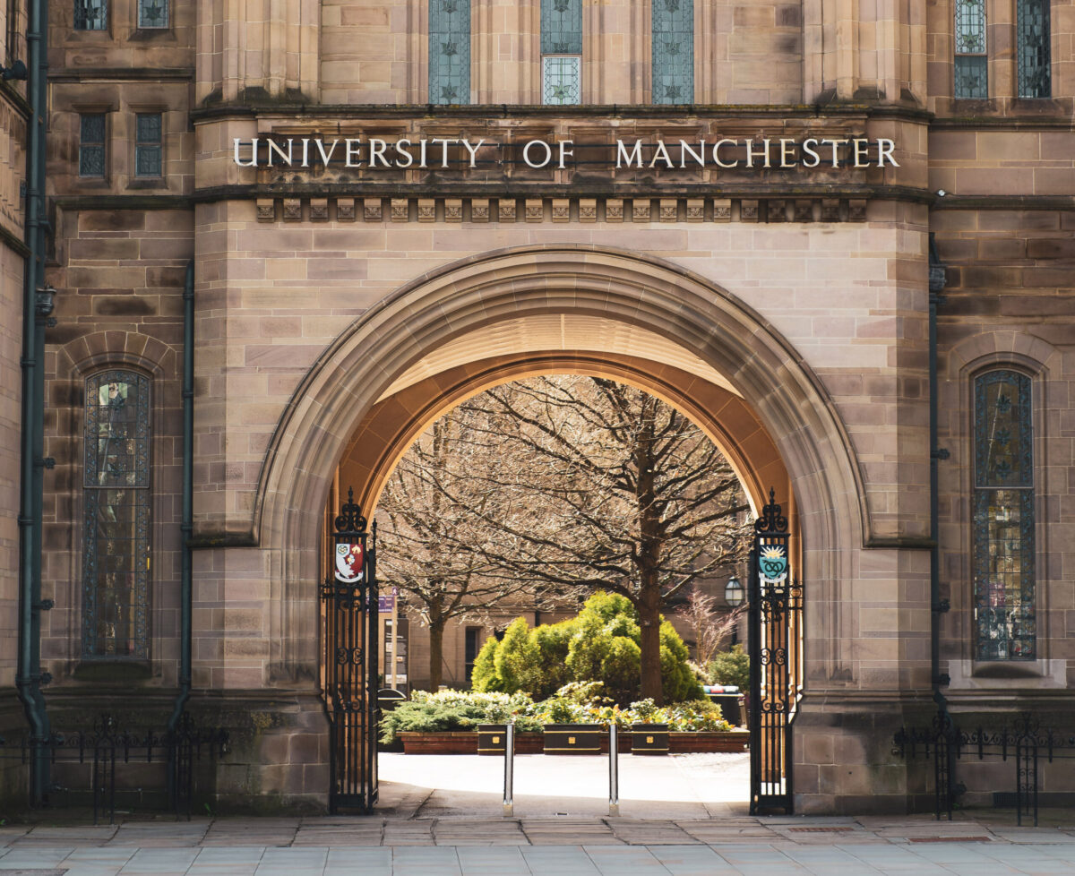 A photo of a stone arch above a wide pavement. A metal sign with the words 'University of Manchester' is fixed on the brickwork above the arch.