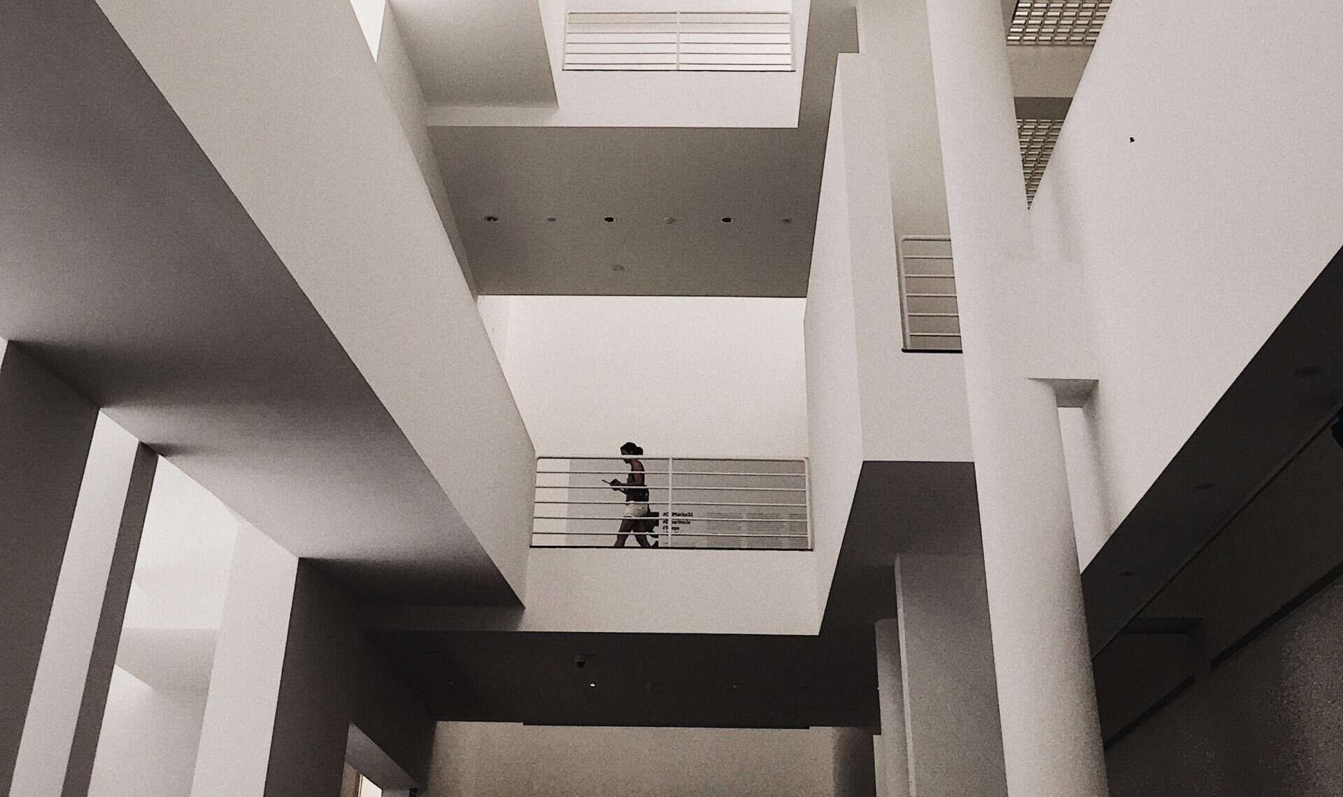 A monochrome photo of a modern building atrium. On the first floor up, a woman is walking over the landing looking at her mobile phone.