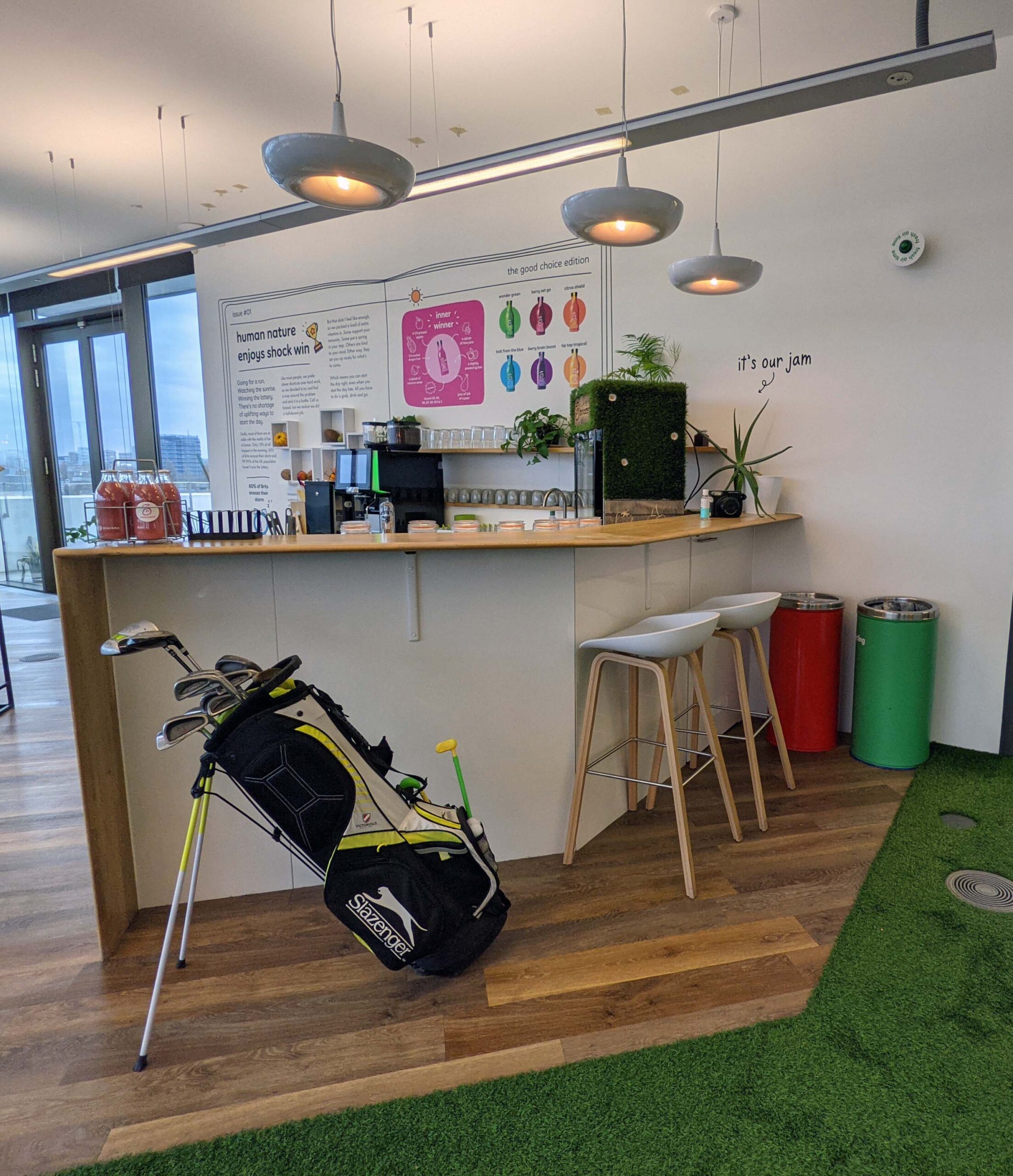 A photo of a colourful office. In the middle of the office is mini kitchen with bottles of smoothies on the sideboard. In front of that is a golf bag. The floor is laid with wood and astroturf.