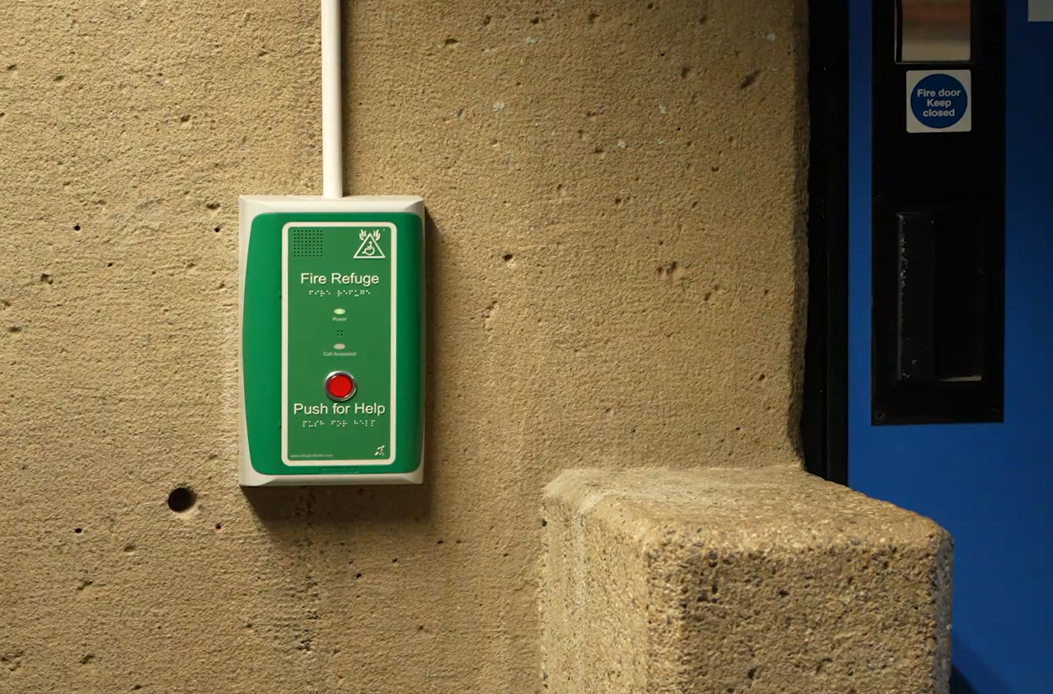 A wireless EVCS outstation is fixed onto a wall, next to a blue door. The words 'Fire Refuge' are written at the top of the outstation. Further down is a red button with the words 'press for help'.